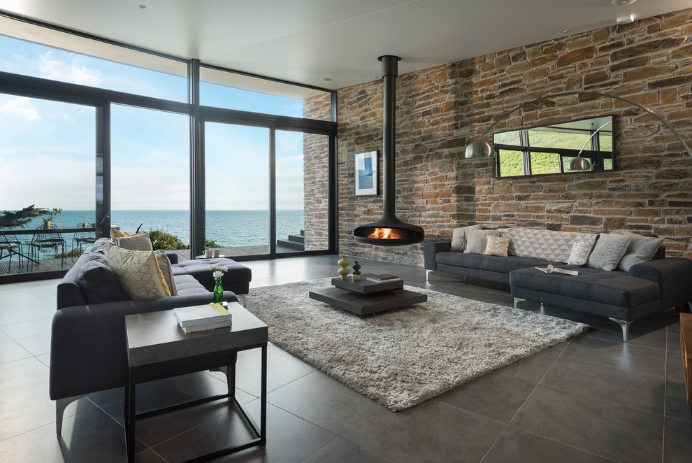 Großes, Offenes Modernes Wohnzimmer mit Porzellan-Bodenfliesen, Hängekamin, grauem Boden und Steinwänden in Cornwall