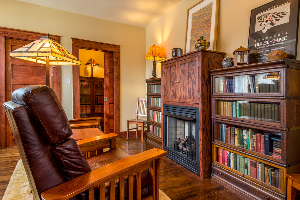 Foto på ett amerikanskt vardagsrum, med ett bibliotek, beige väggar, mörkt trägolv, en standard öppen spis och en spiselkrans i tegelsten