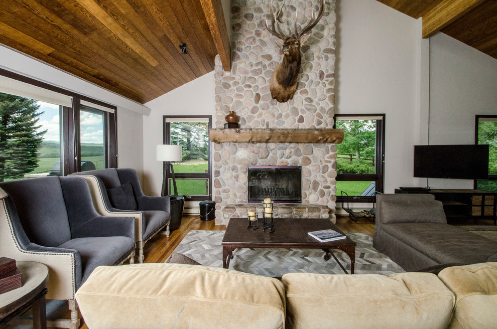 Imagen de salón abierto rural grande con paredes blancas, suelo de madera en tonos medios, todas las chimeneas, marco de chimenea de piedra y televisor colgado en la pared