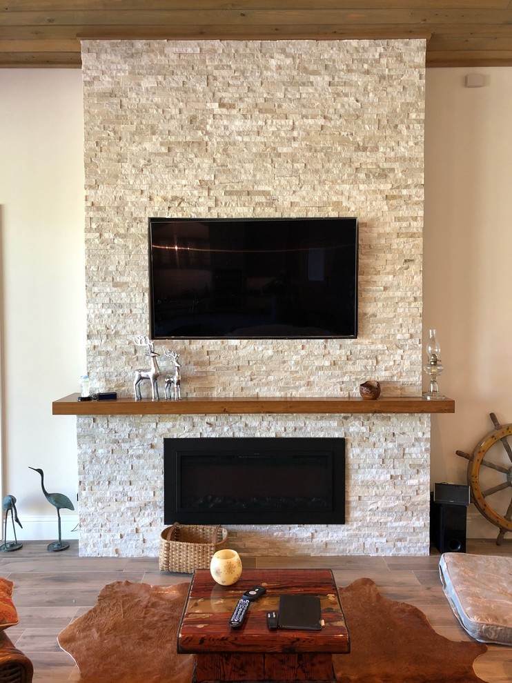 На фото: большая открытая гостиная комната в стиле кантри с серыми стенами, полом из керамогранита, горизонтальным камином, фасадом камина из камня, телевизором на стене и коричневым полом