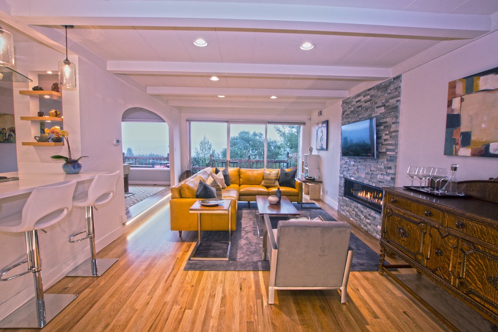 Cette image montre un grand salon minimaliste ouvert avec un mur blanc, un sol en bois brun, une cheminée ribbon, un manteau de cheminée en pierre et un téléviseur fixé au mur.