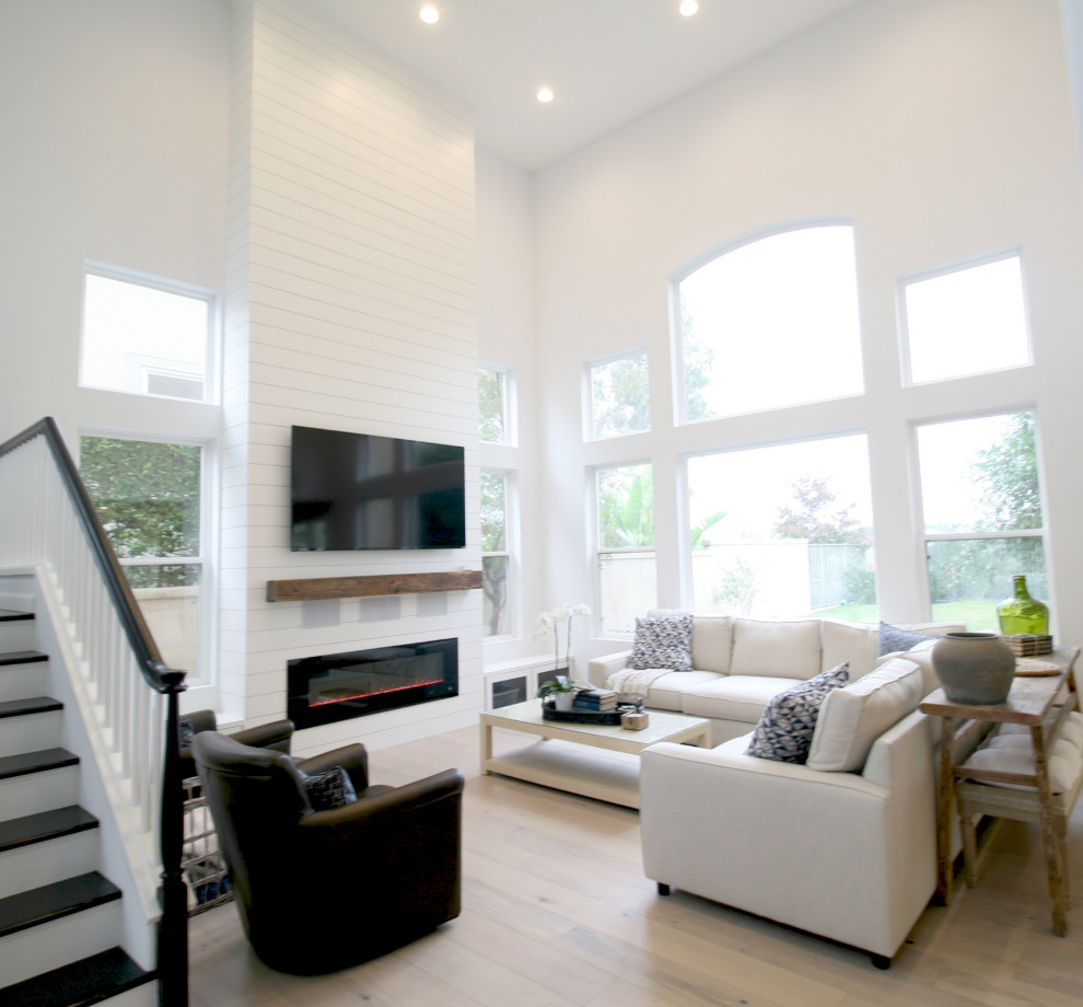 Modelo de salón abierto de estilo de casa de campo de tamaño medio con paredes blancas, suelo de madera clara, chimenea lineal, marco de chimenea de madera, televisor colgado en la pared y suelo beige