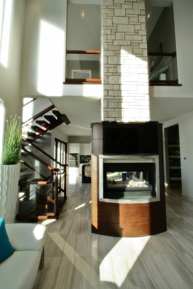 На фото: большая открытая гостиная комната в стиле модернизм с серыми стенами, полом из керамогранита, двусторонним камином и фасадом камина из дерева