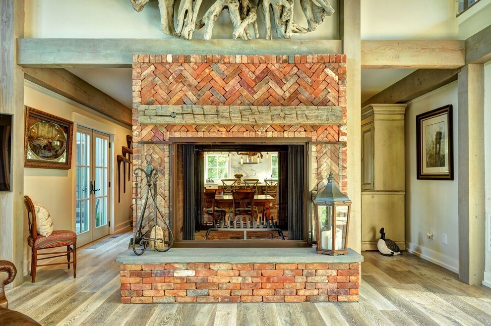 Modelo de salón abierto de estilo de casa de campo grande con paredes beige, suelo de madera clara, chimenea de doble cara y marco de chimenea de ladrillo