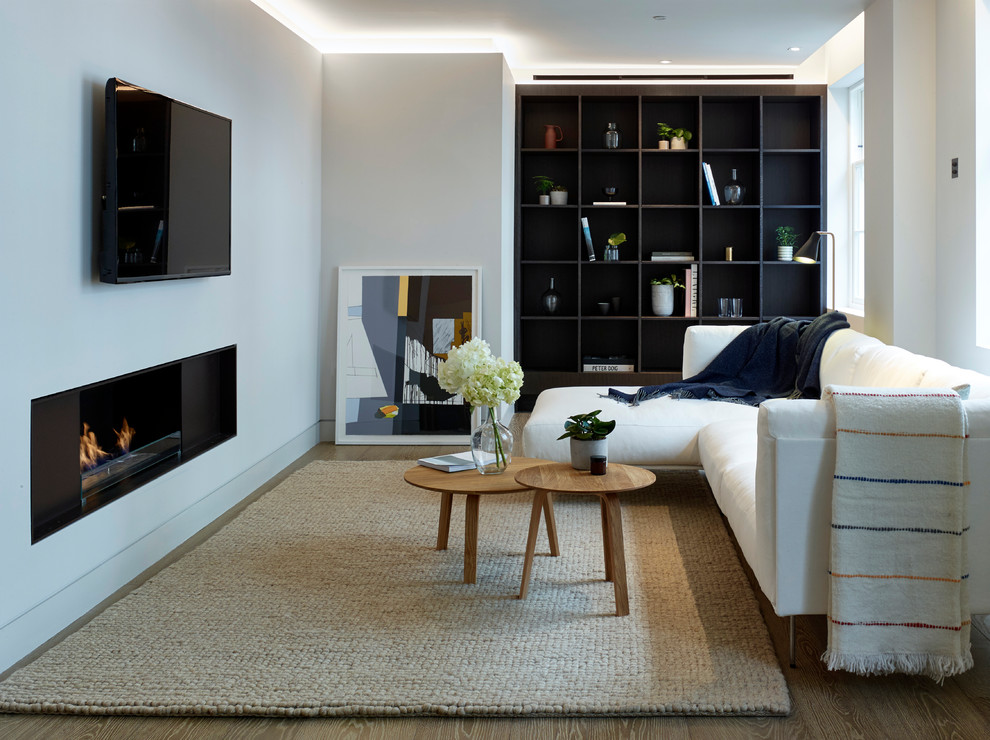 На фото: открытая гостиная комната среднего размера в современном стиле с с книжными шкафами и полками, серыми стенами, светлым паркетным полом, горизонтальным камином, фасадом камина из металла и телевизором на стене с
