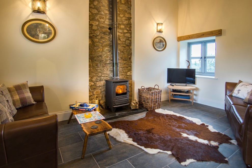 Foto de salón abierto campestre de tamaño medio con paredes beige, suelo de pizarra, estufa de leña, marco de chimenea de piedra y televisor independiente