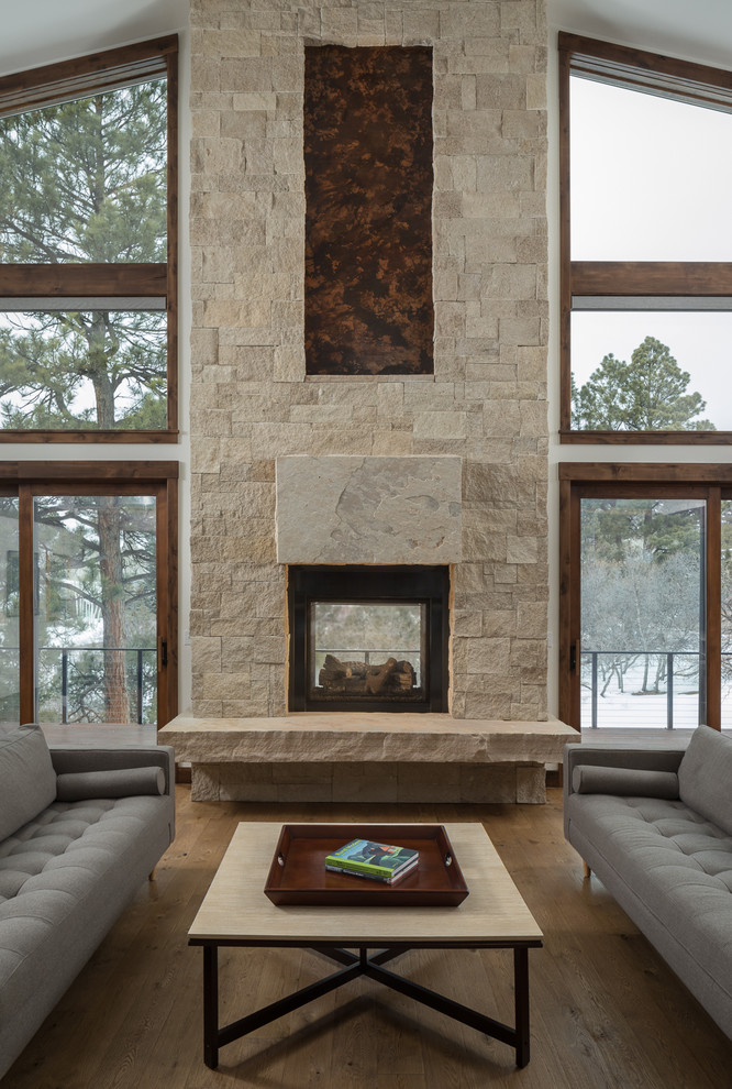 Modelo de salón rústico con paredes blancas, suelo de madera en tonos medios, chimenea de doble cara, marco de chimenea de piedra y suelo marrón