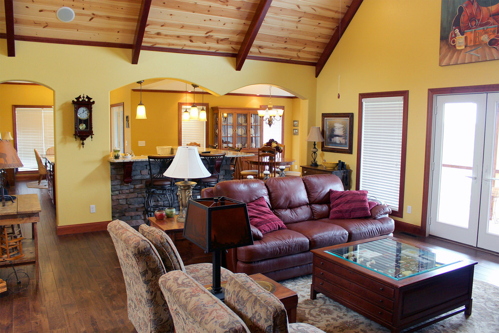 На фото: большая открытая гостиная комната в стиле рустика с желтыми стенами, темным паркетным полом, стандартным камином и фасадом камина из камня без телевизора
