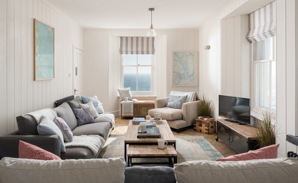 Modelo de salón cerrado costero de tamaño medio con paredes blancas, suelo de madera clara, televisor independiente y suelo beige