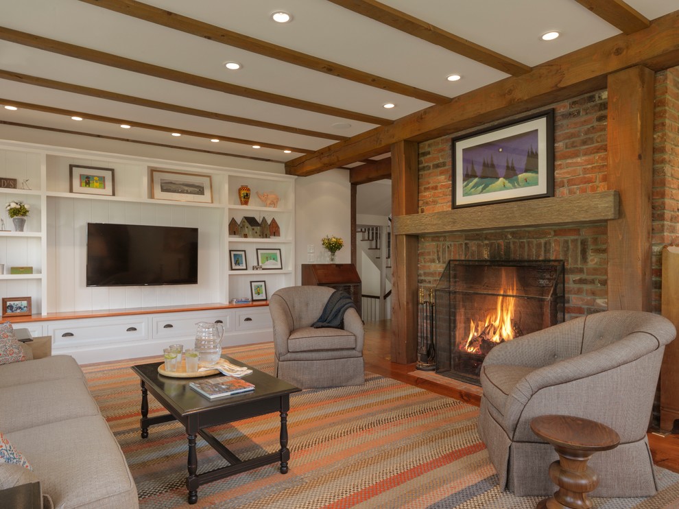 Bild på ett lantligt vardagsrum, med vita väggar, mellanmörkt trägolv, en standard öppen spis, en spiselkrans i tegelsten, en väggmonterad TV och brunt golv