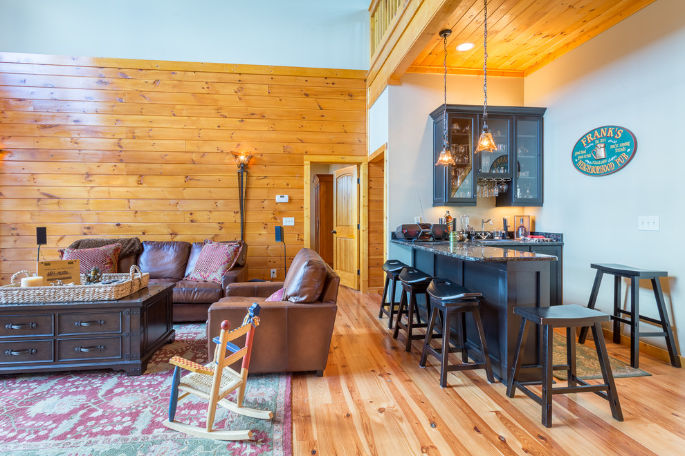 Ejemplo de salón con barra de bar tipo loft rural grande con paredes beige, suelo de madera en tonos medios, chimenea de doble cara, marco de chimenea de piedra y televisor colgado en la pared