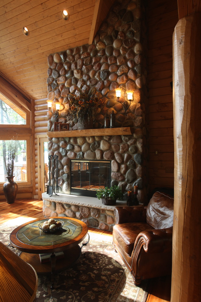 Modelo de salón abierto rural grande sin televisor con paredes marrones, suelo de madera en tonos medios, todas las chimeneas y marco de chimenea de piedra