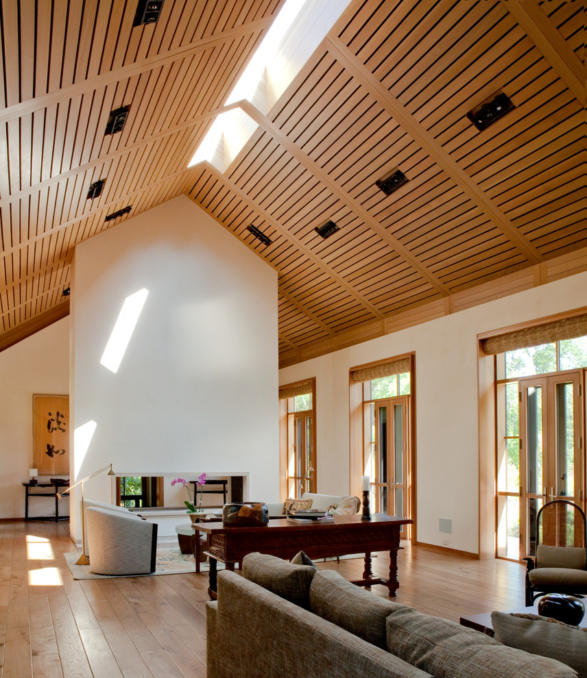 Diseño de salón tradicional renovado grande con paredes blancas, suelo de madera clara, chimenea de doble cara y marco de chimenea de piedra
