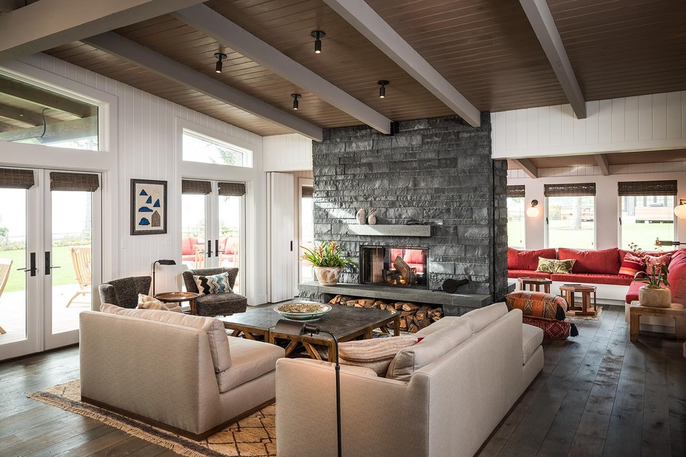 Coastal open plan living room in New York with white walls, dark hardwood flooring, a two-sided fireplace, a stone fireplace surround and brown floors.