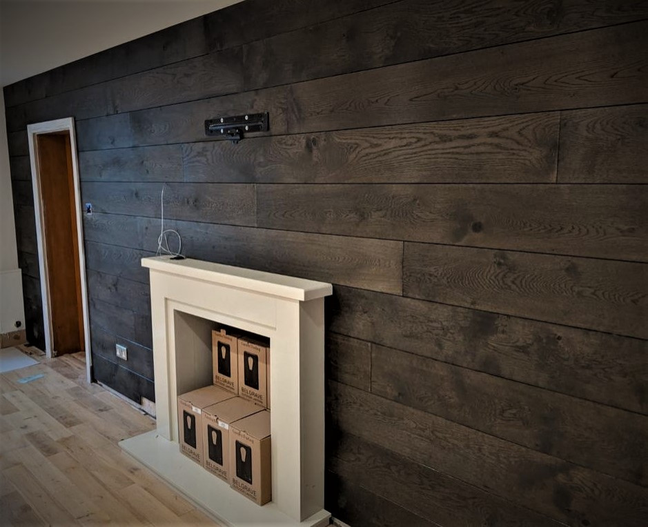 This is an example of a large rustic open plan living room in Hertfordshire with brown walls, light hardwood flooring, a wooden fireplace surround, a wall mounted tv and brown floors.