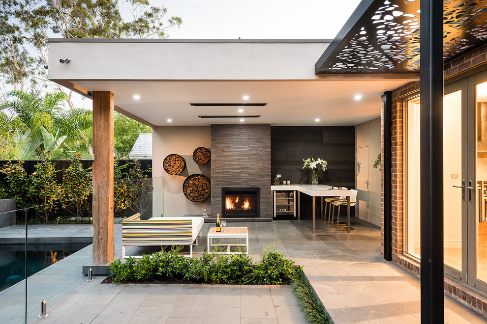 Réalisation d'une grande terrasse arrière minimaliste avec un foyer extérieur, des pavés en béton et une extension de toiture.