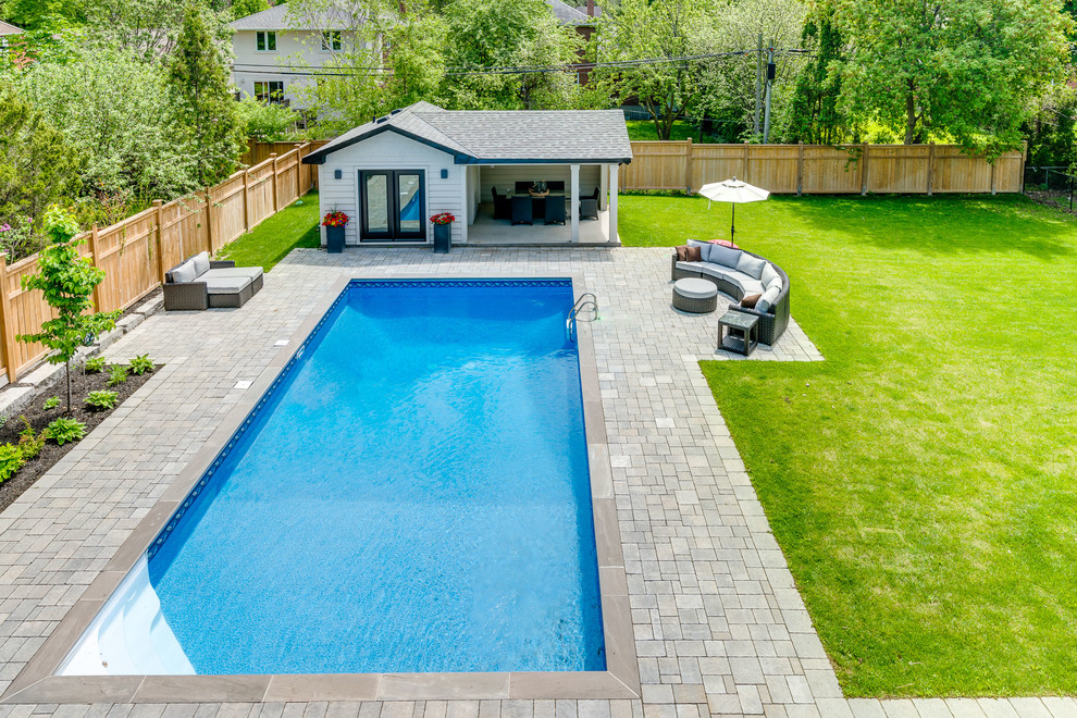 Стильный дизайн: большой прямоугольный, спортивный бассейн на заднем дворе в стиле неоклассика (современная классика) с домиком у бассейна и покрытием из каменной брусчатки - последний тренд