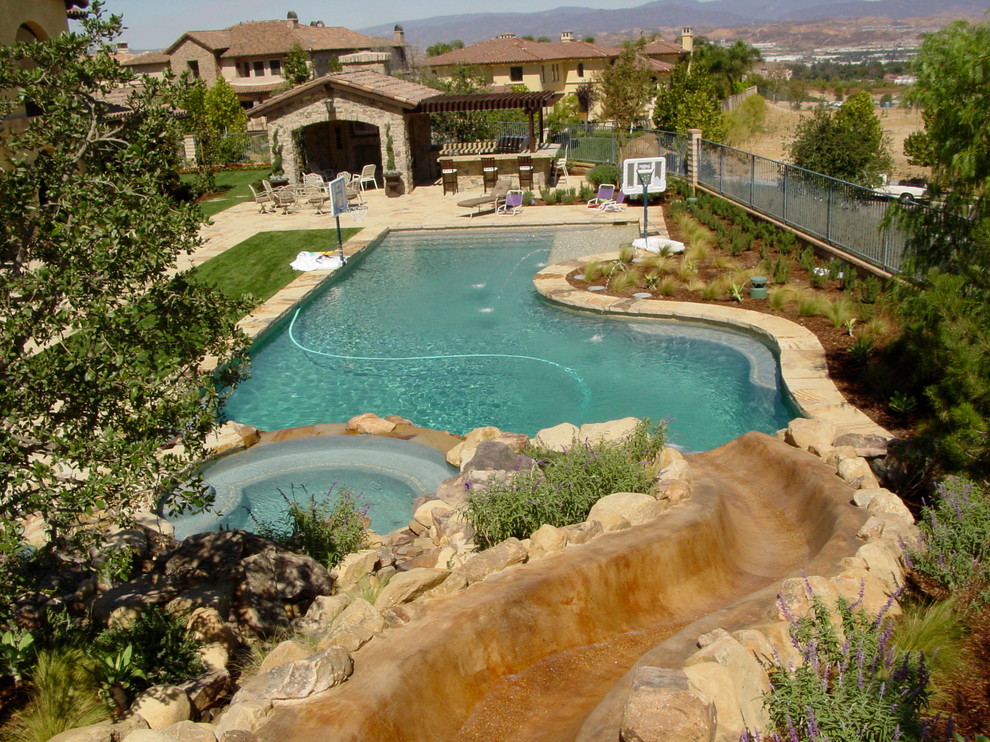 Expansive world-inspired back custom shaped lengths swimming pool in Los Angeles with a water slide and natural stone paving.