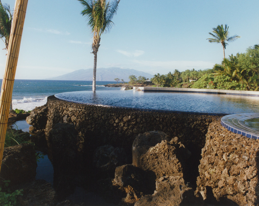 Foto di un'ampia piscina a sfioro infinito tropicale rotonda dietro casa con un acquascivolo e pavimentazioni in pietra naturale