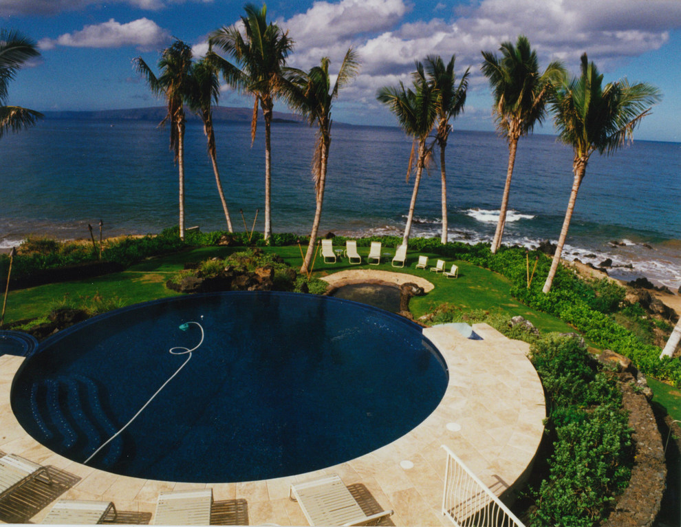 Idee per un'ampia piscina a sfioro infinito tropicale rotonda dietro casa con un acquascivolo e pavimentazioni in pietra naturale