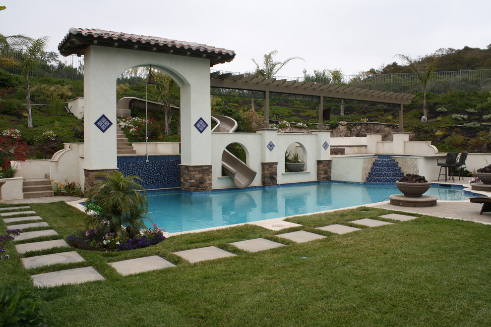 Imagen de piscina con tobogán alargada mediterránea grande a medida en patio trasero con adoquines de piedra natural