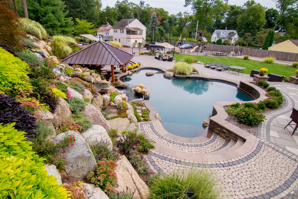 На фото: огромный естественный бассейн произвольной формы на заднем дворе с джакузи и мощением тротуарной плиткой с