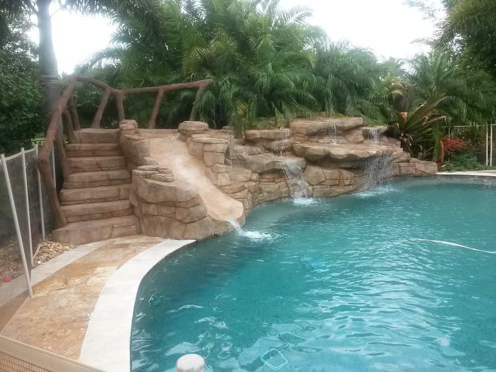 Large world-inspired back custom shaped natural swimming pool in Miami with a water slide and natural stone paving.
