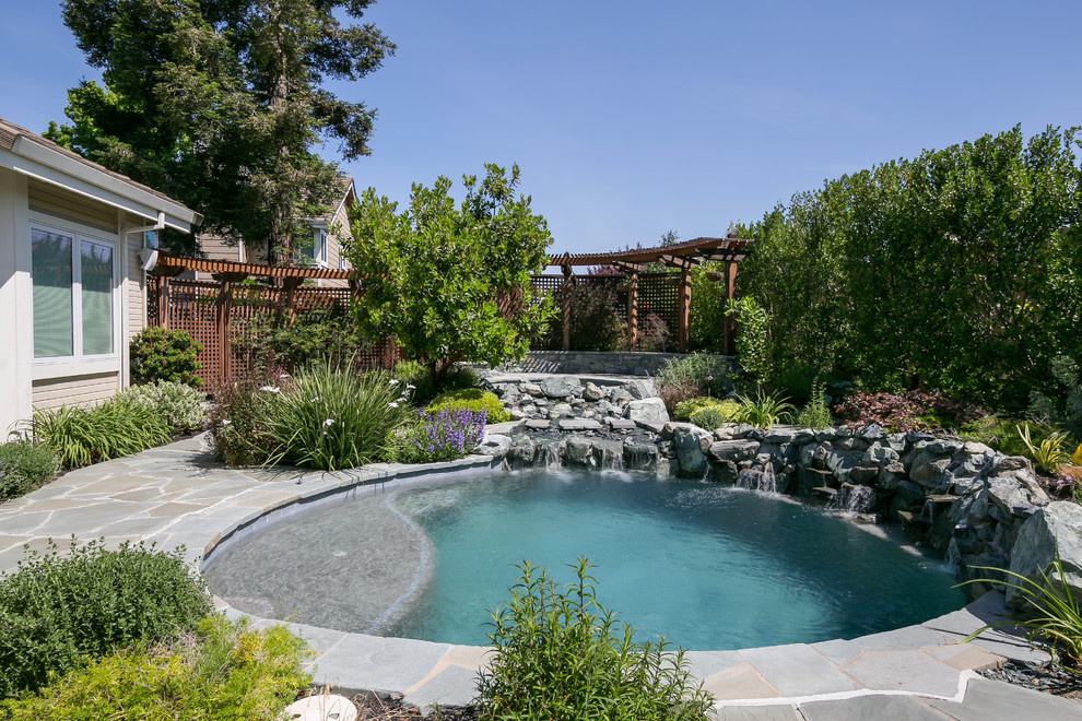 Modelo de piscina con fuente natural tropical pequeña redondeada en patio trasero con adoquines de piedra natural