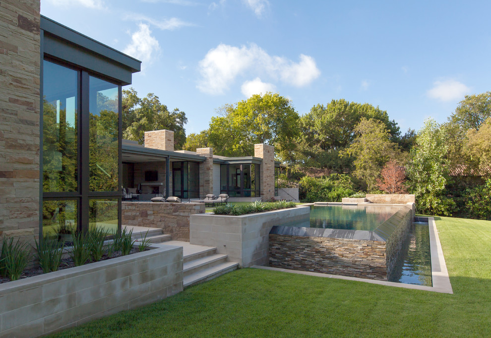 This is an example of a medium sized modern back rectangular infinity swimming pool in Dallas with a water feature and natural stone paving.