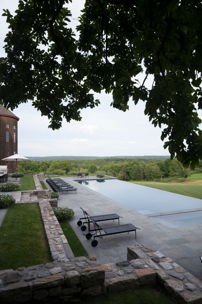 Large country backyard stone and rectangular infinity pool photo in New York
