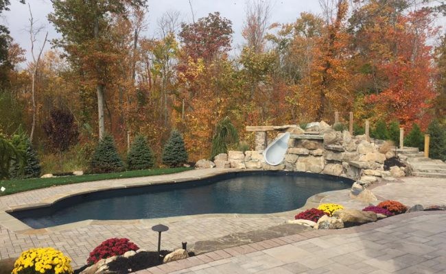 Large classic back custom shaped lengths swimming pool in New York with a water slide and brick paving.