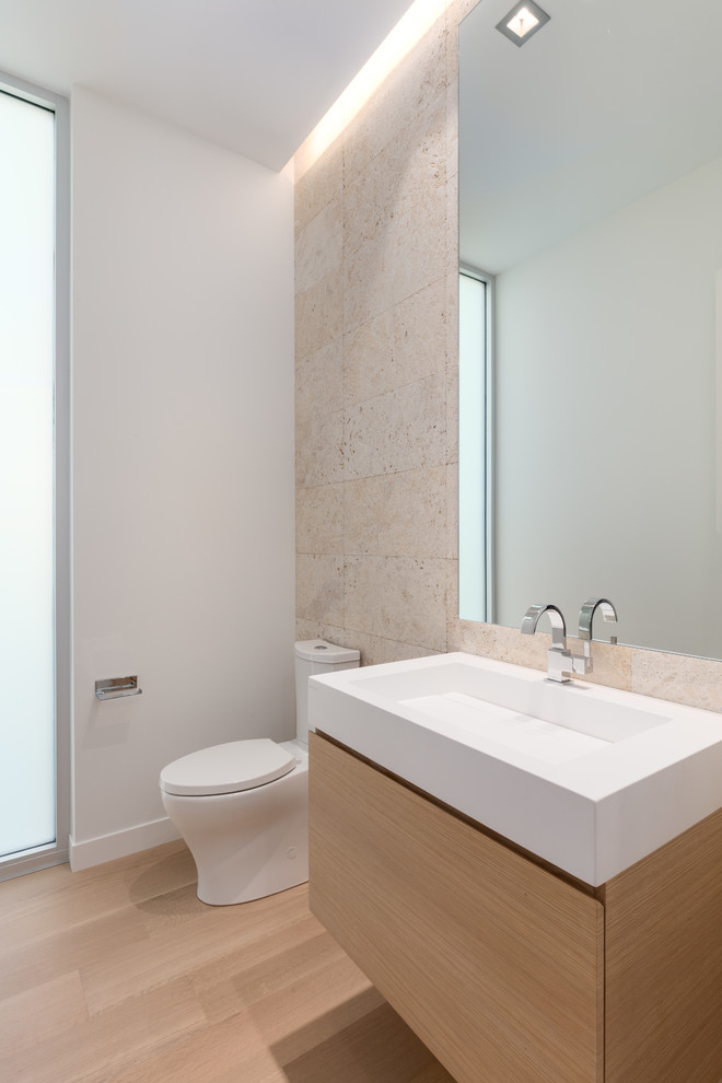 Inspiration for a mid-sized contemporary beige tile and limestone tile light wood floor powder room remodel in Tampa with flat-panel cabinets, light wood cabinets, a one-piece toilet, white walls, an integrated sink and quartz countertops
