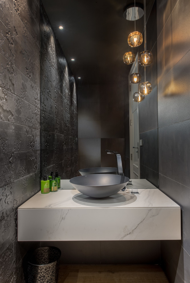 Inspiration for a large contemporary gray tile medium tone wood floor and brown floor powder room remodel in Auckland with beige cabinets, a one-piece toilet, a vessel sink, flat-panel cabinets, black walls and tile countertops