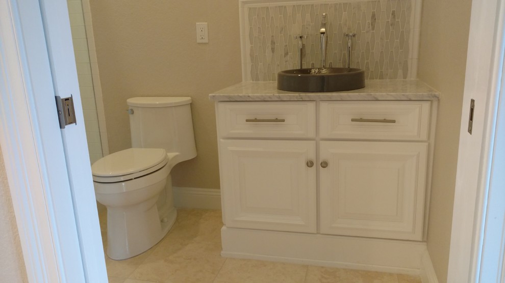 This is an example of a medium sized beach style cloakroom in Miami with raised-panel cabinets, white cabinets, a two-piece toilet, beige tiles, stone tiles, white walls, travertine flooring, a vessel sink and marble worktops.