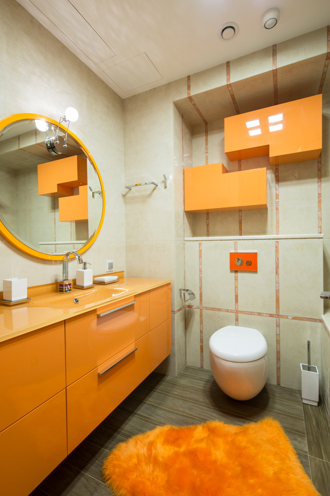 Ejemplo de aseo contemporáneo con armarios con paneles lisos, puertas de armario naranjas, encimera de vidrio, sanitario de pared, baldosas y/o azulejos beige y encimeras naranjas