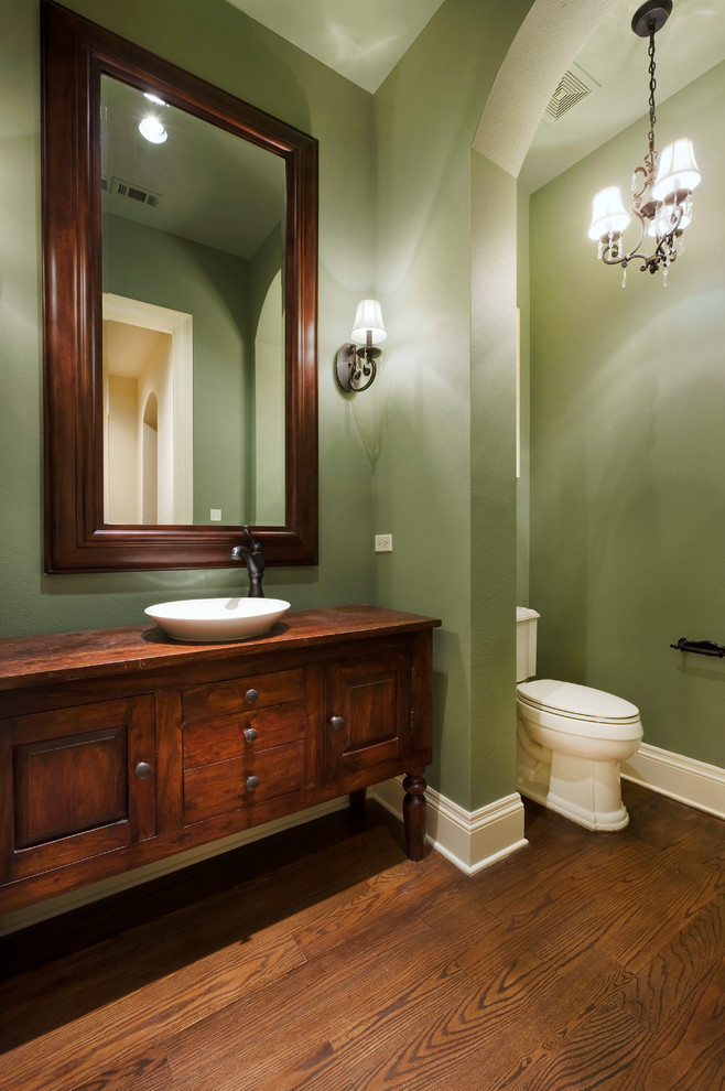 This is an example of a traditional cloakroom in Austin with freestanding cabinets, dark wood cabinets, a two-piece toilet, green walls, medium hardwood flooring, a vessel sink, wooden worktops and brown worktops.