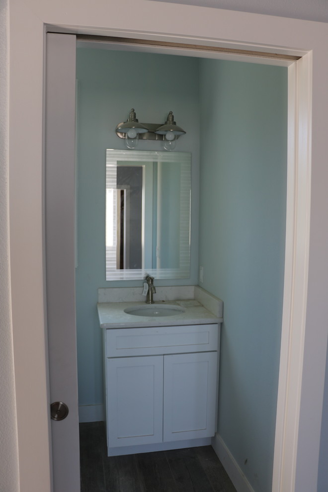 Cette image montre un WC et toilettes marin de taille moyenne avec un placard à porte shaker, des portes de placard blanches, un mur bleu, parquet foncé, un lavabo encastré, un plan de toilette en quartz et un sol marron.
