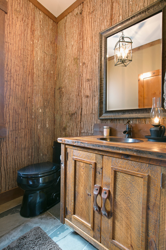 Cette image montre un grand WC et toilettes chalet en bois brun avec un lavabo posé, un plan de toilette en bois, WC à poser, un sol en ardoise et un placard avec porte à panneau encastré.