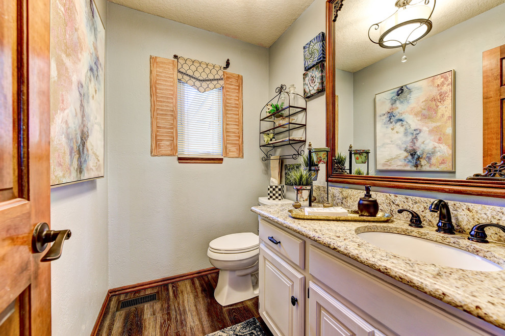 Aménagement d'un WC et toilettes classique de taille moyenne avec un placard avec porte à panneau surélevé, des portes de placard blanches, WC séparés, un mur bleu, un sol en bois brun, un lavabo encastré, un plan de toilette en granite et un plan de toilette beige.
