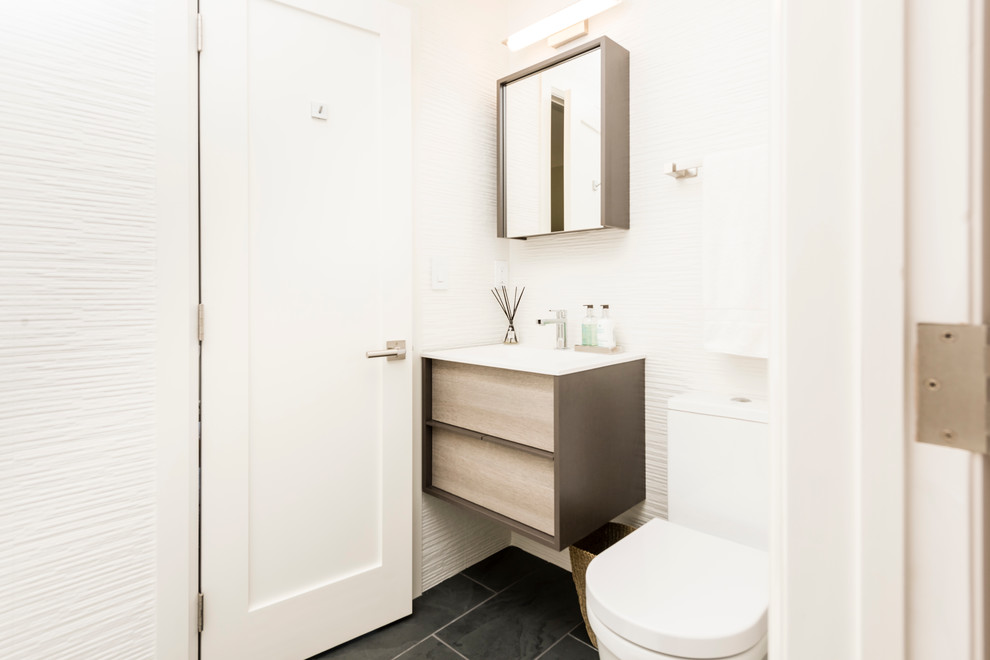 Design ideas for a medium sized contemporary cloakroom in New York with light wood cabinets, a one-piece toilet, white tiles, white walls, ceramic flooring, a built-in sink, solid surface worktops, black floors, flat-panel cabinets, stone tiles and white worktops.