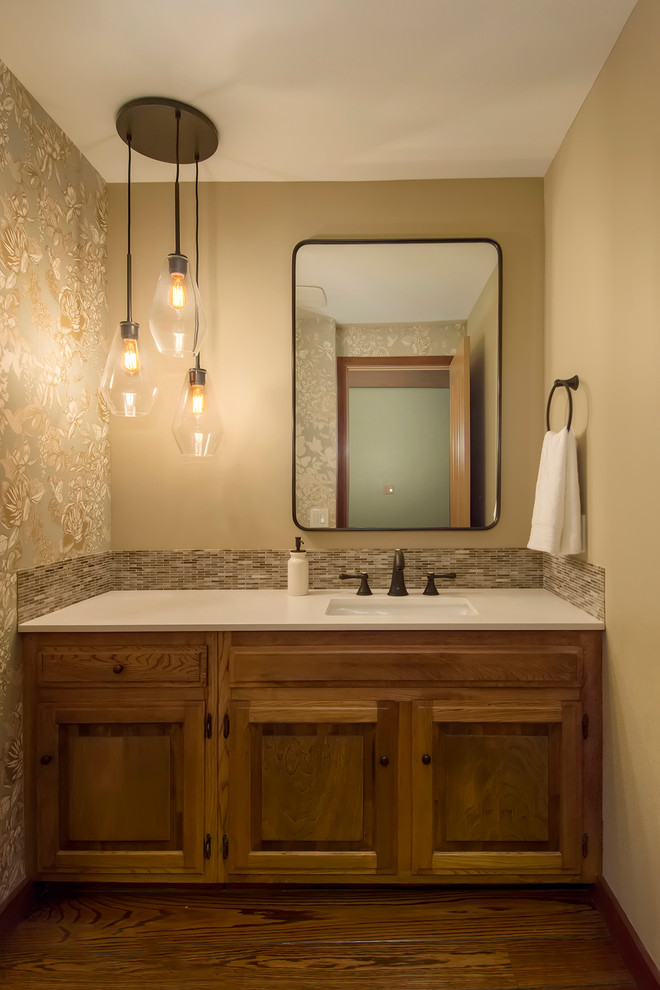 This is an example of a small classic cloakroom in Seattle with raised-panel cabinets, brown cabinets, a wall mounted toilet, multi-coloured tiles, glass tiles, beige walls, medium hardwood flooring, a submerged sink, engineered stone worktops, brown floors and white worktops.