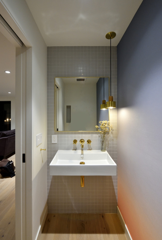 Example of a mid-sized minimalist gray tile and ceramic tile light wood floor and beige floor powder room design in New York with a wall-mount toilet, gray walls, a wall-mount sink and solid surface countertops