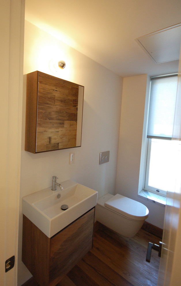 Medium sized scandinavian cloakroom in New York with flat-panel cabinets, medium wood cabinets, a wall mounted toilet, white walls, dark hardwood flooring, a wall-mounted sink and brown floors.