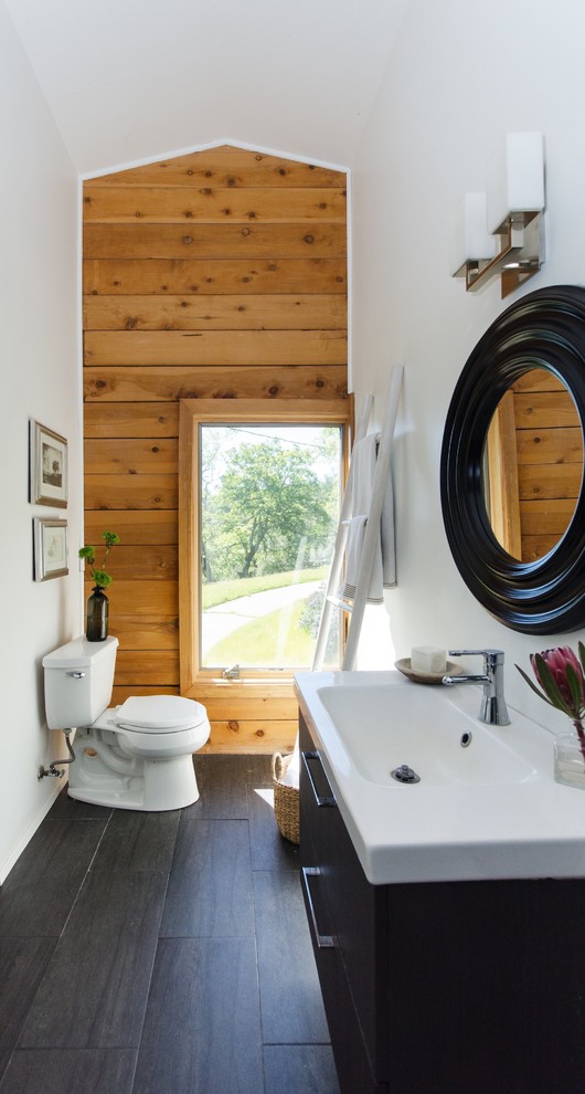 Modelo de aseo de estilo de casa de campo con sanitario de dos piezas, paredes blancas, lavabo integrado, armarios con paneles lisos, puertas de armario negras y suelo gris