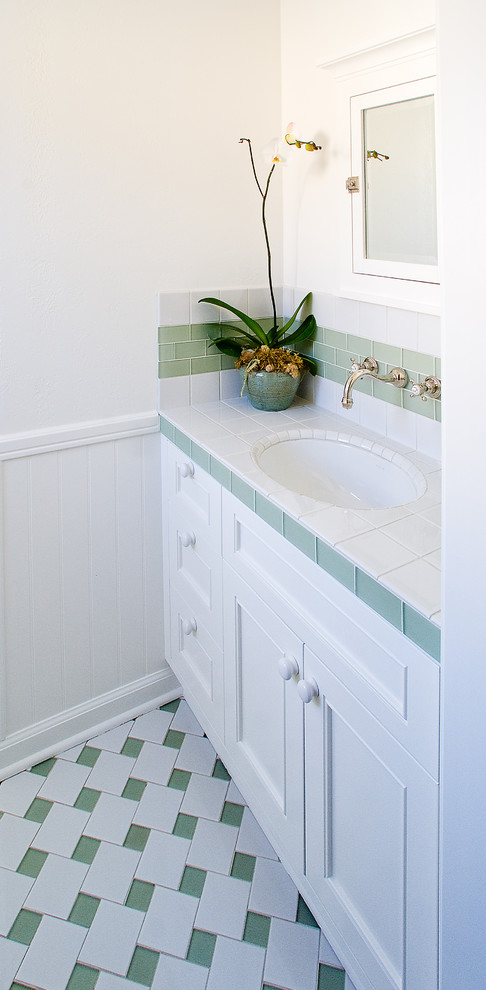 Klassisk inredning av ett litet toalett, med ett undermonterad handfat, vita skåp, kaklad bänkskiva, vit kakel, grön kakel, glaskakel, vita väggar, klinkergolv i porslin och luckor med infälld panel