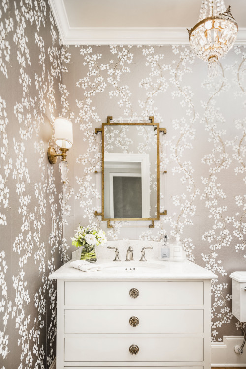 The small antique chandelier was the inspiration for this powder room, while the acrylic in the wall sconce and mirror add a touch of updated transitional style. The metallic grey wallpaper contrasts beautifully with the white vanity cabinet. The quartz counter top has subtle grey veining with a shaped backsplash and ogee edge and offers a nice backdrop for the polished nickel faucet. Brass on the mirror and sconce tie in nicely with the chinoiserie corner shelf, while framed fern art fill the need for just one piece of art. The powder room offers a space of elegance, if only for a moment!
Inspiration for a transitional medium tone wood floor and wallpaper powder room remodel in Charlotte with white cabinets, an undermount sink, quartz countertops, white countertops, flat-panel cabinets, gray walls and a freestanding vanity