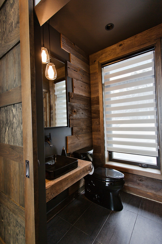 Kleine Urige Gästetoilette mit schwarzen Fliesen, schwarzer Wandfarbe, Keramikboden, Aufsatzwaschbecken, Waschtisch aus Holz und brauner Waschtischplatte in Montreal