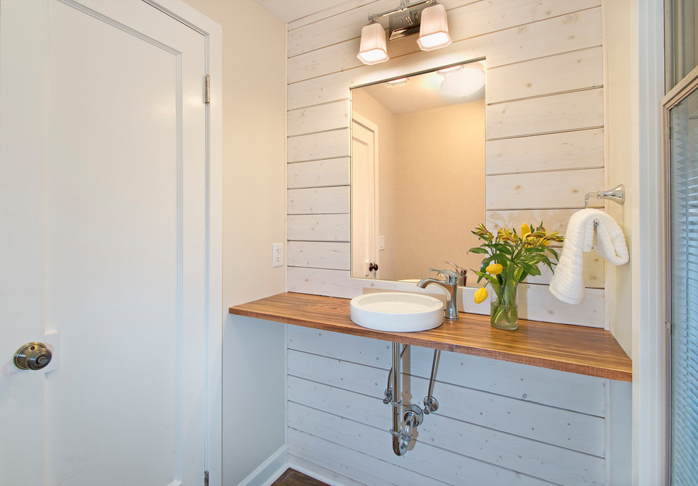 Modelo de aseo campestre pequeño con sanitario de una pieza, paredes blancas, suelo de madera en tonos medios, lavabo sobreencimera y encimera de madera