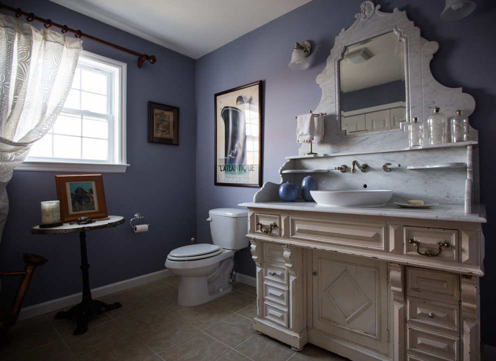 Example of a mid-sized eclectic beige tile and ceramic tile powder room design in New York with a vessel sink, raised-panel cabinets, white cabinets, marble countertops and a two-piece toilet