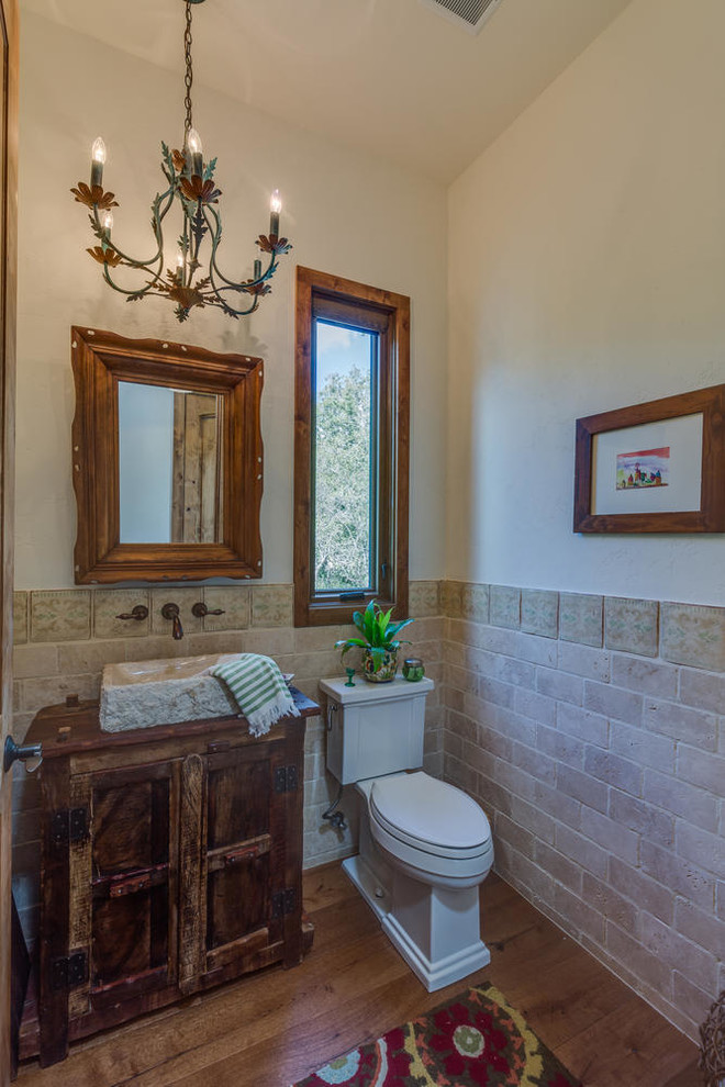 Rustik inredning av ett mellanstort toalett, med möbel-liknande, bruna skåp, en toalettstol med separat cisternkåpa, beige kakel, stenkakel, vita väggar, ljust trägolv, ett fristående handfat, träbänkskiva och brunt golv